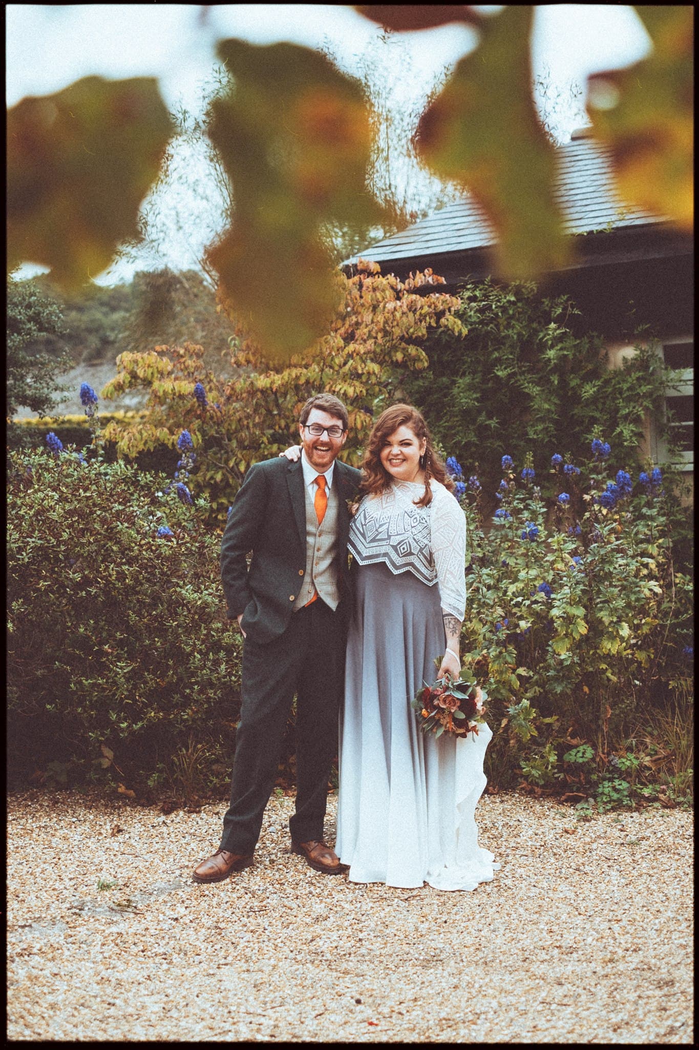 A couple in wedding attire standing in a garden with greenery and flowers.