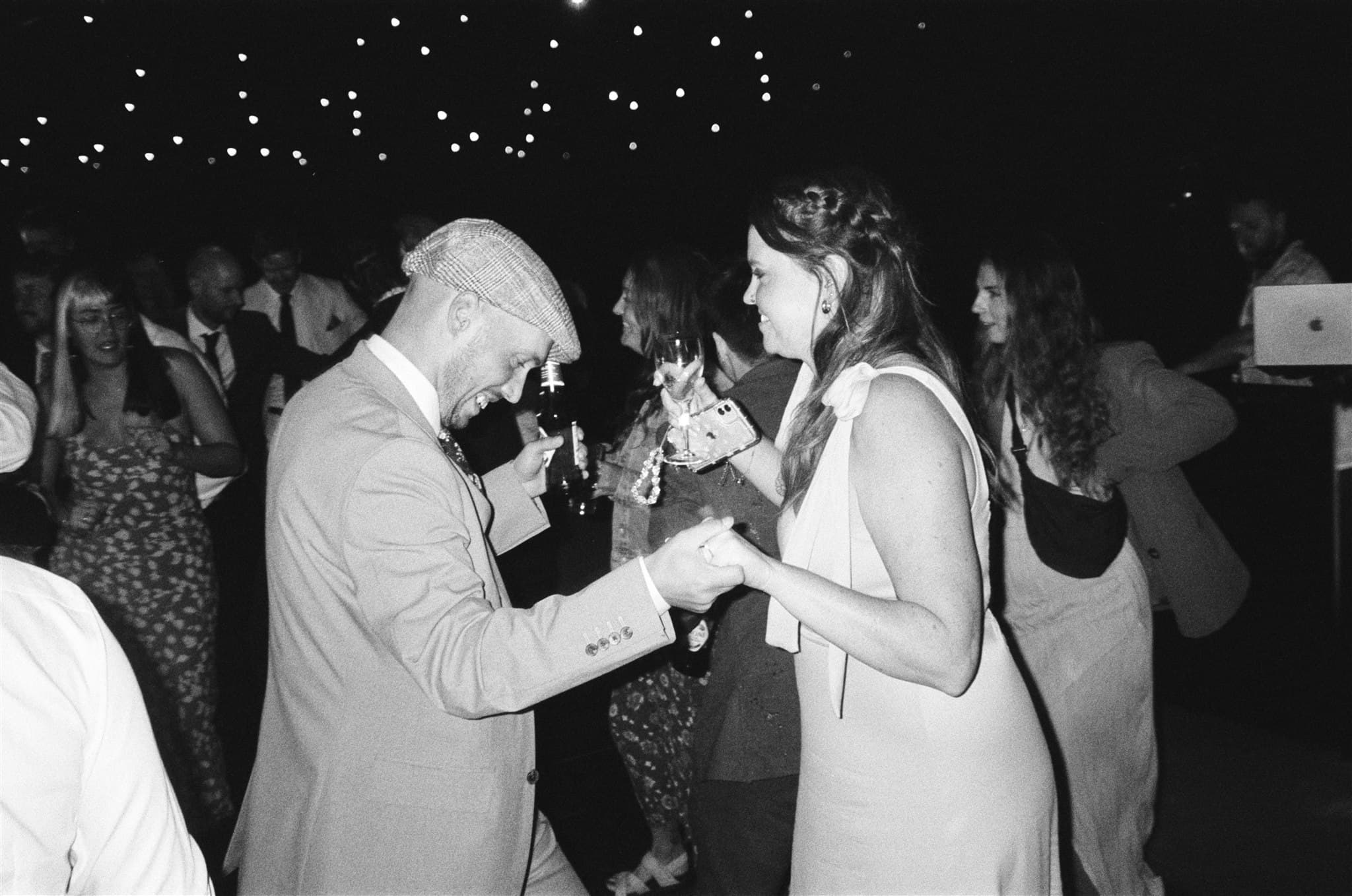Film photo, Black and white image of people at a social event, some holding drinks, with string lights overhead.