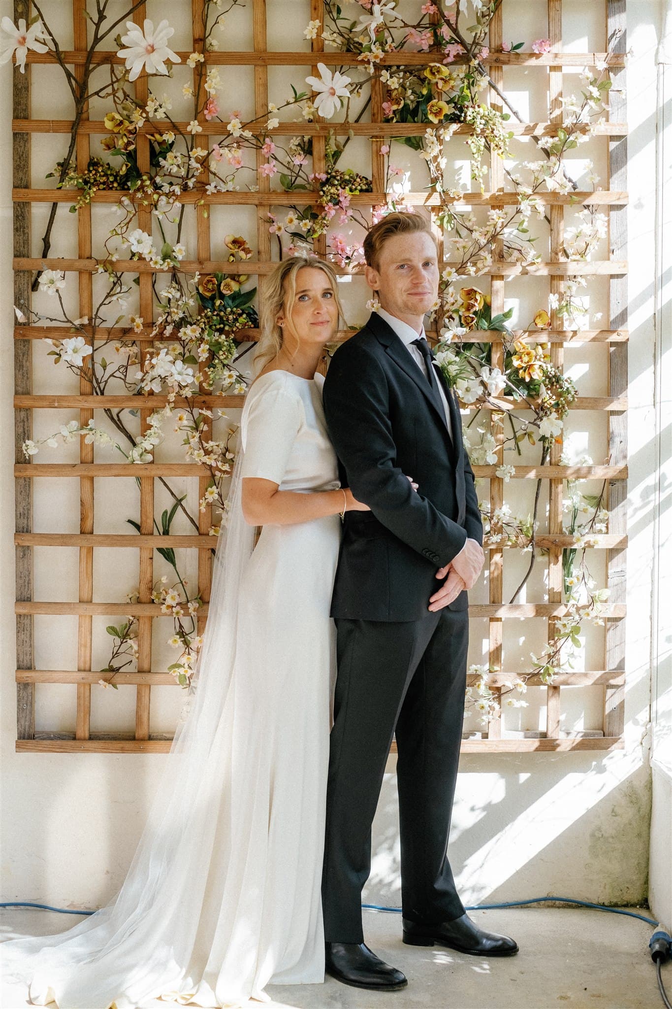 A couple in wedding attire standing against a floral backdrop.