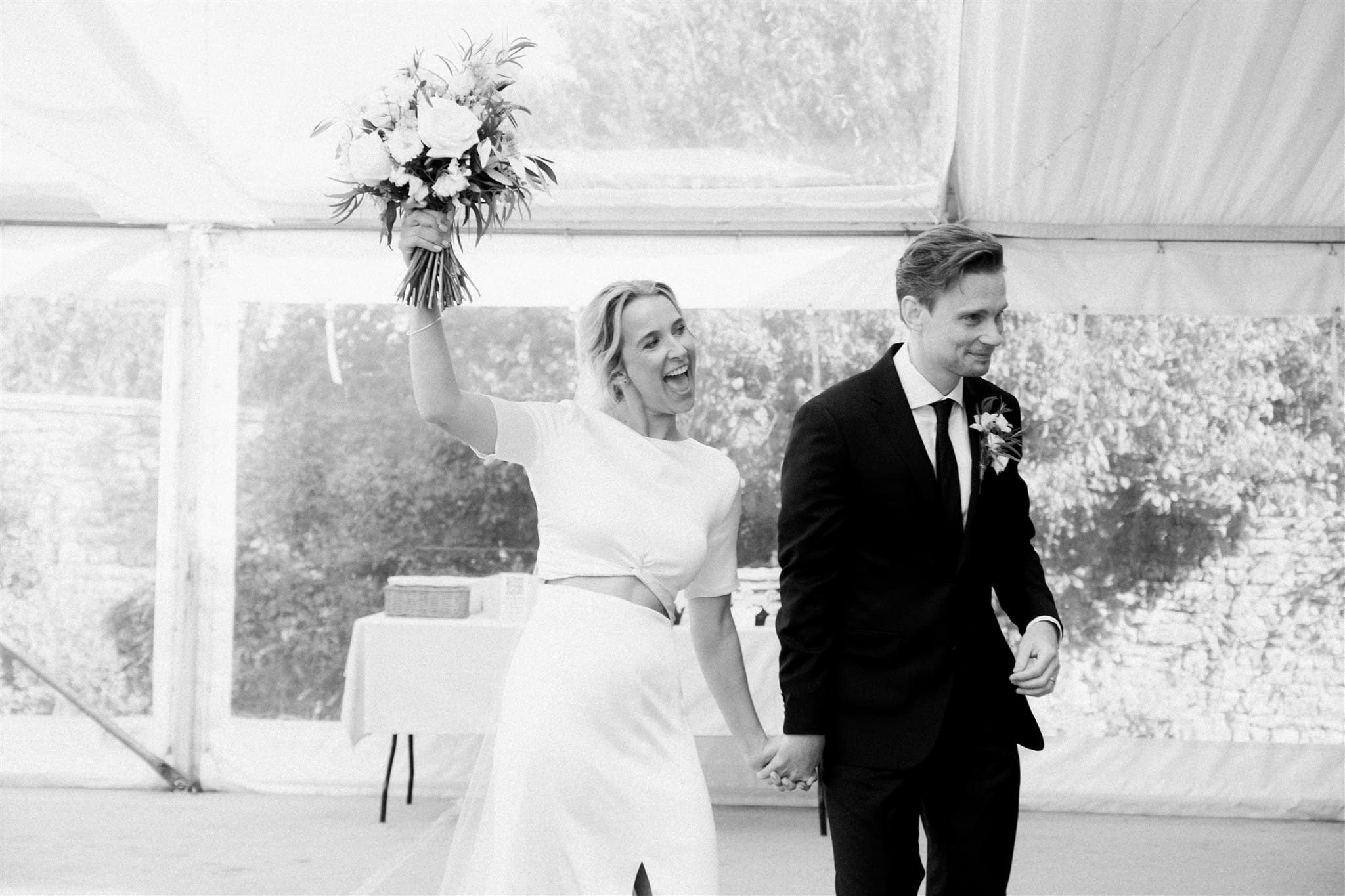 Bride in white raising bouquet next to groom in black suit at wedding.
