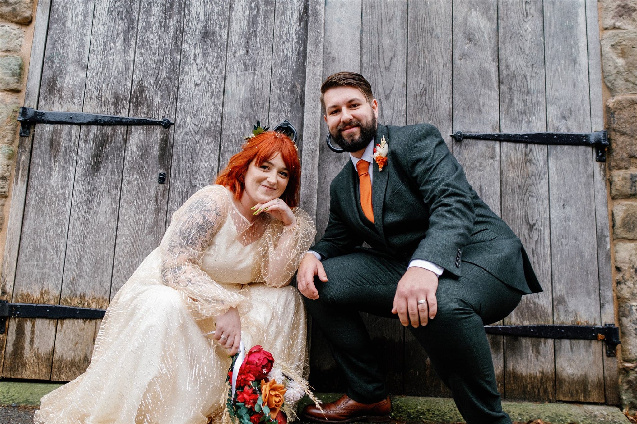 A bride in a glittery dress and a groom in a suit sitting in front of a wooden door.