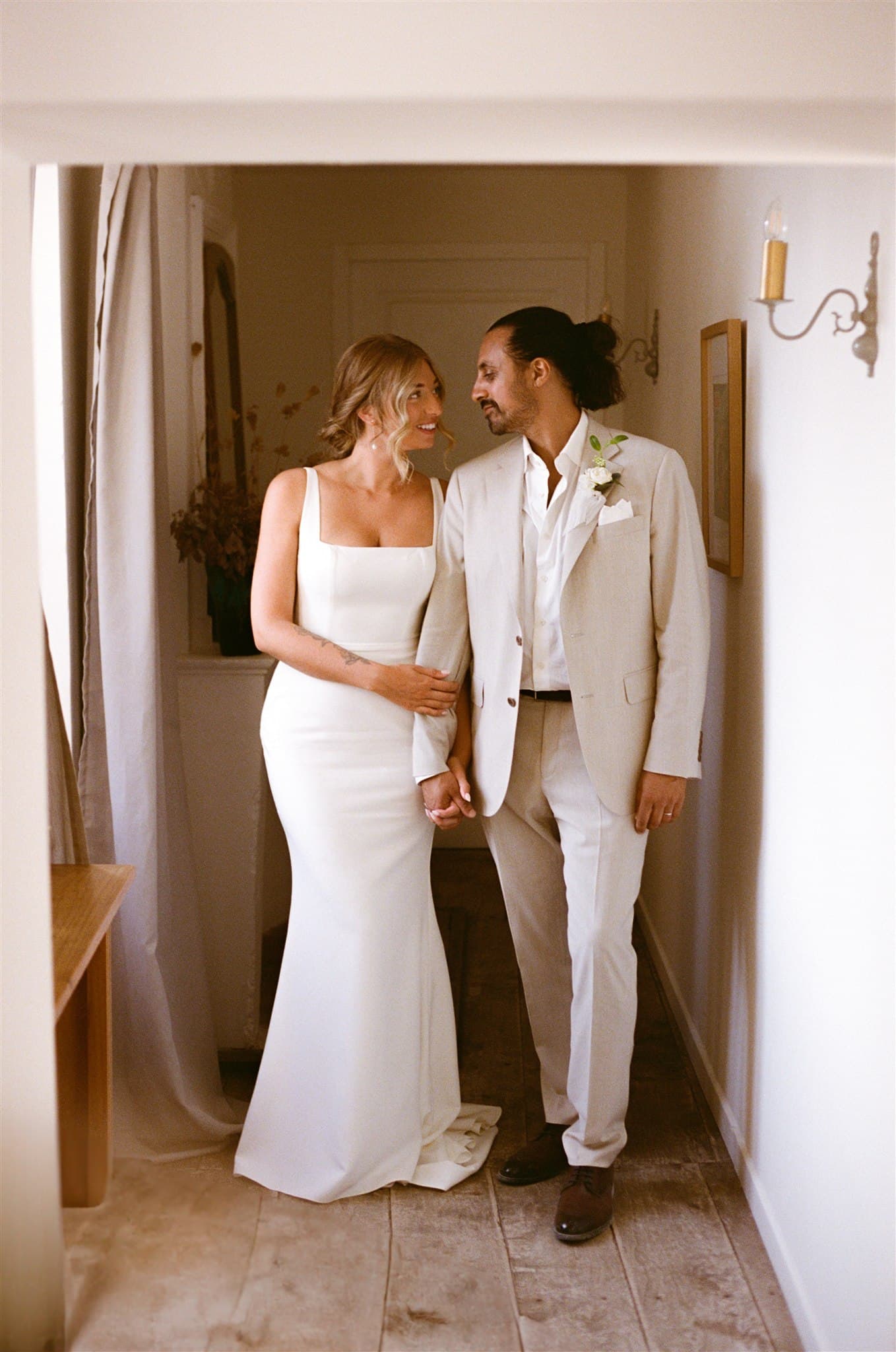 A couple in wedding attire stands in a warmly lit hallway, holding hands and smiling at each other. The bride wears a white dress and the groom a light suit.