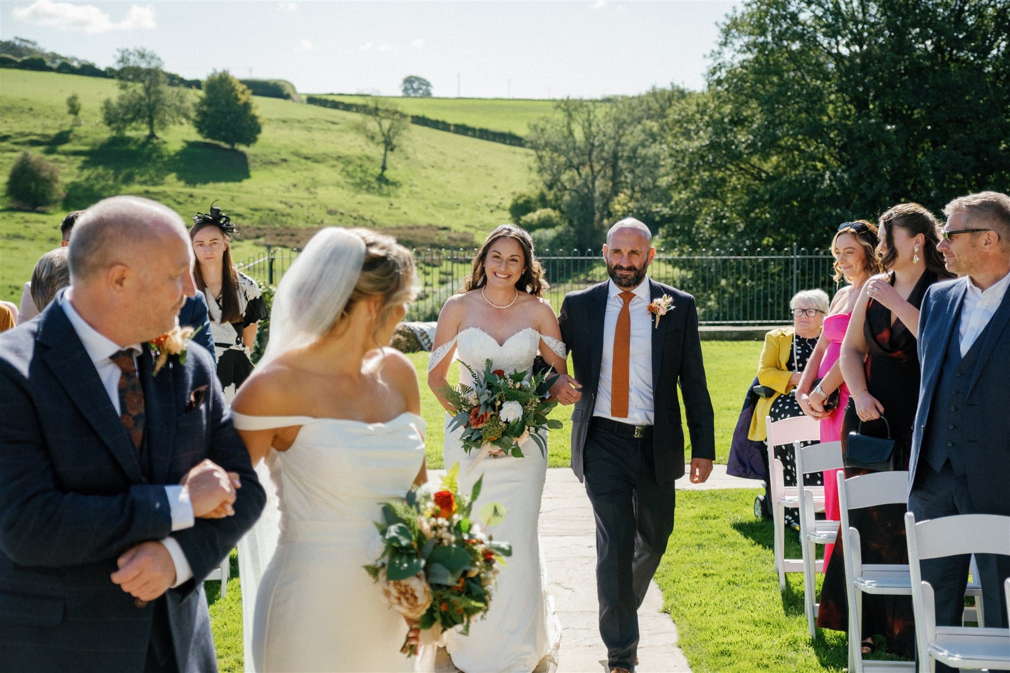 Bride walks down an outdoor aisle, holding vibrant flowers beside a smiling man. Her wife to be smiling back down the aisle at her. Guests watch, seated on a sunny day with green hills in the background.
