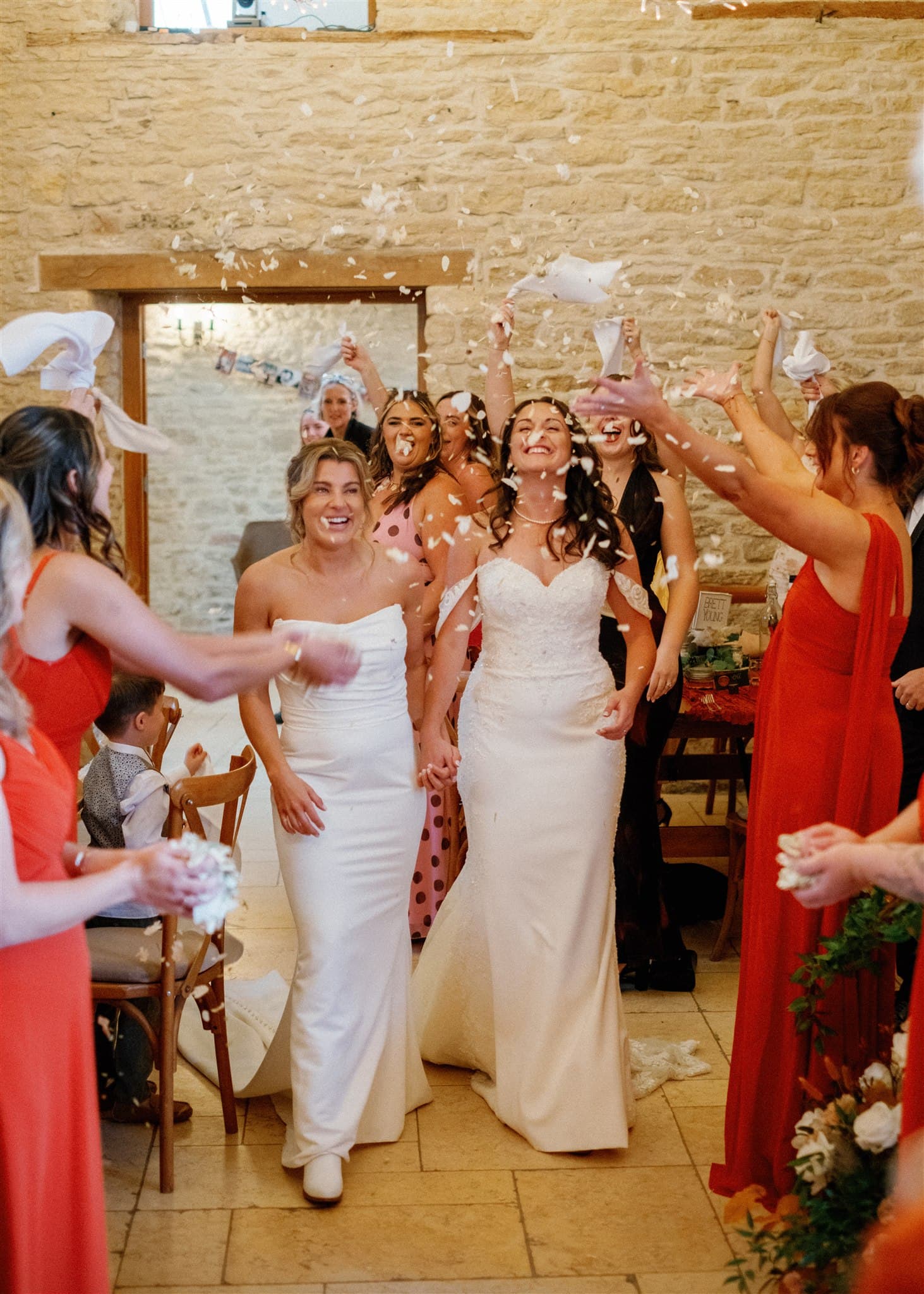 Two brides in white dresses joyfully walk hand in hand down an aisle, surrounded by guests in colorful attire throwing confetti in celebration.