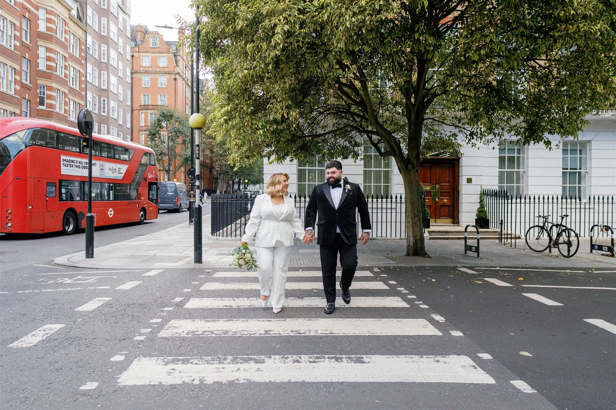 A bride in a white suit and a groom in a tuxedo holding hands, crossing a street.