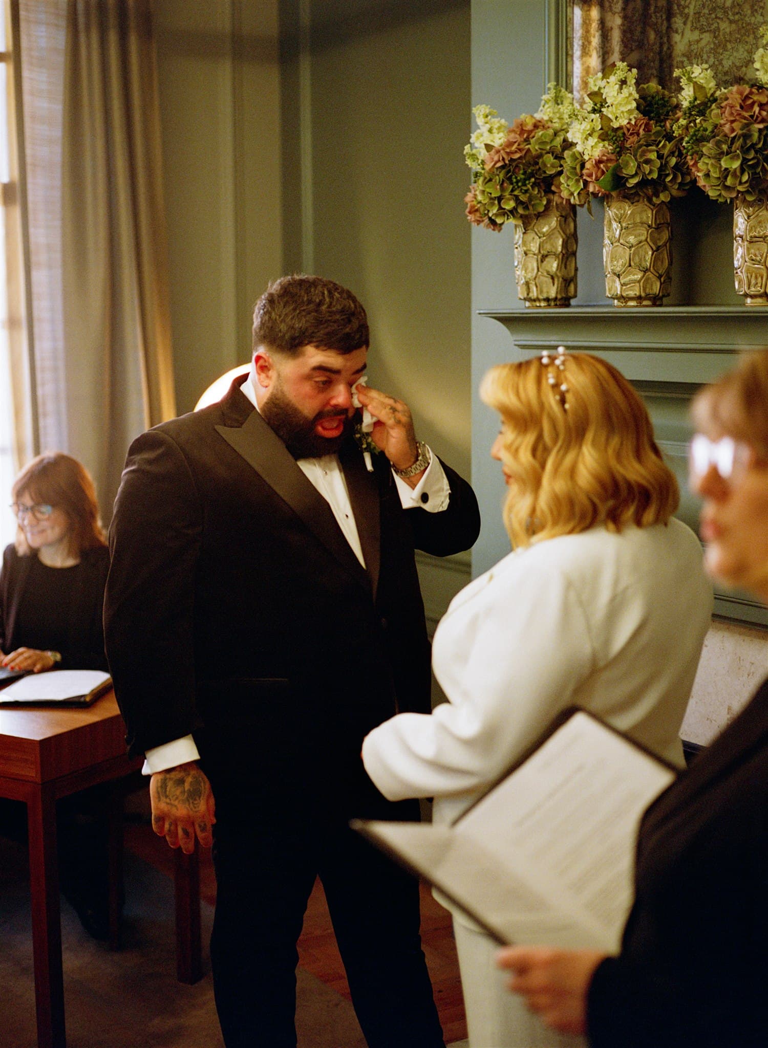 Elegant interior with individuals in formal wear, decorative urns on mantelpiece.
