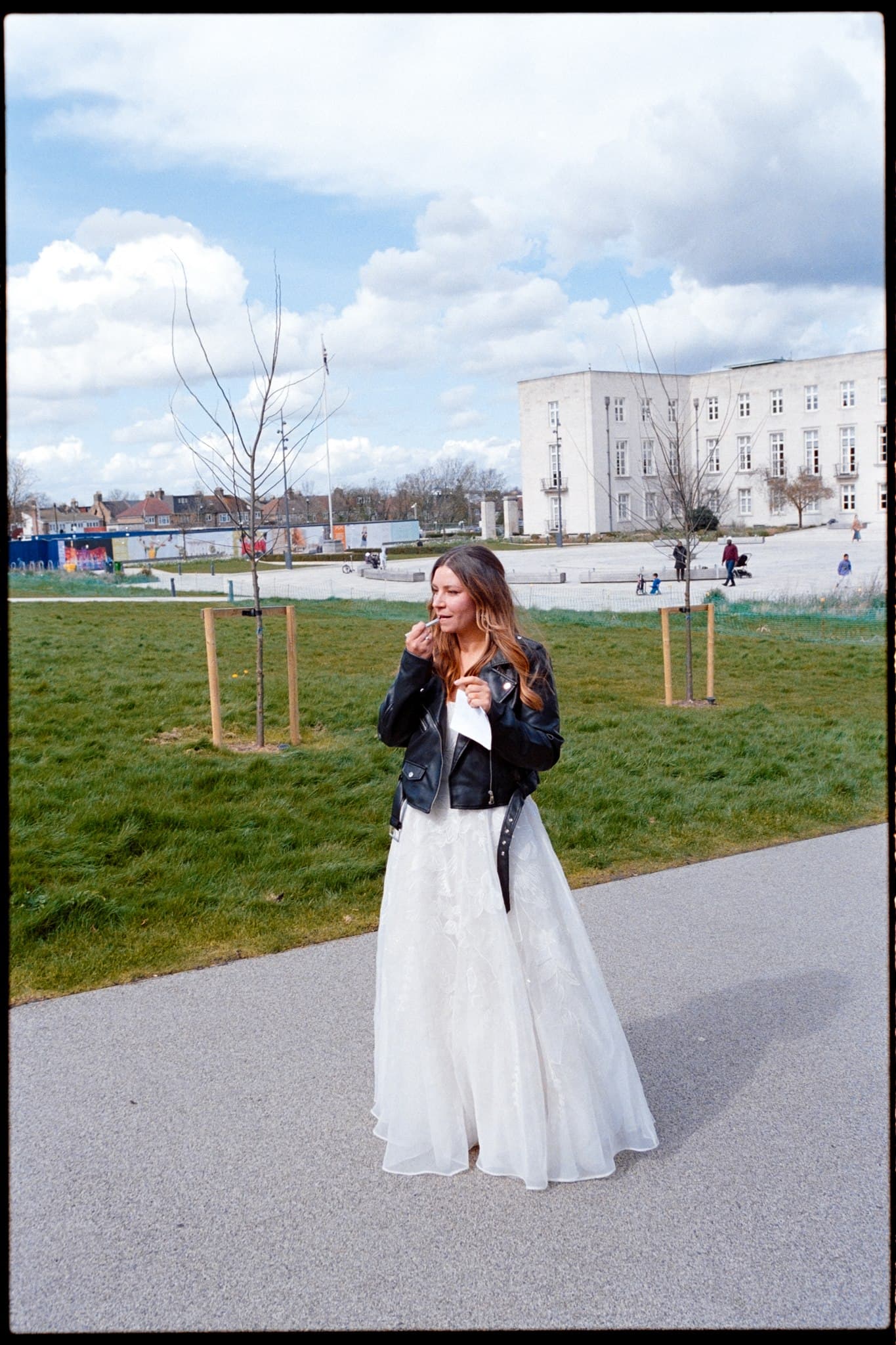 Bride in a white dress and black jacket standing on a path in a park. Film Photo