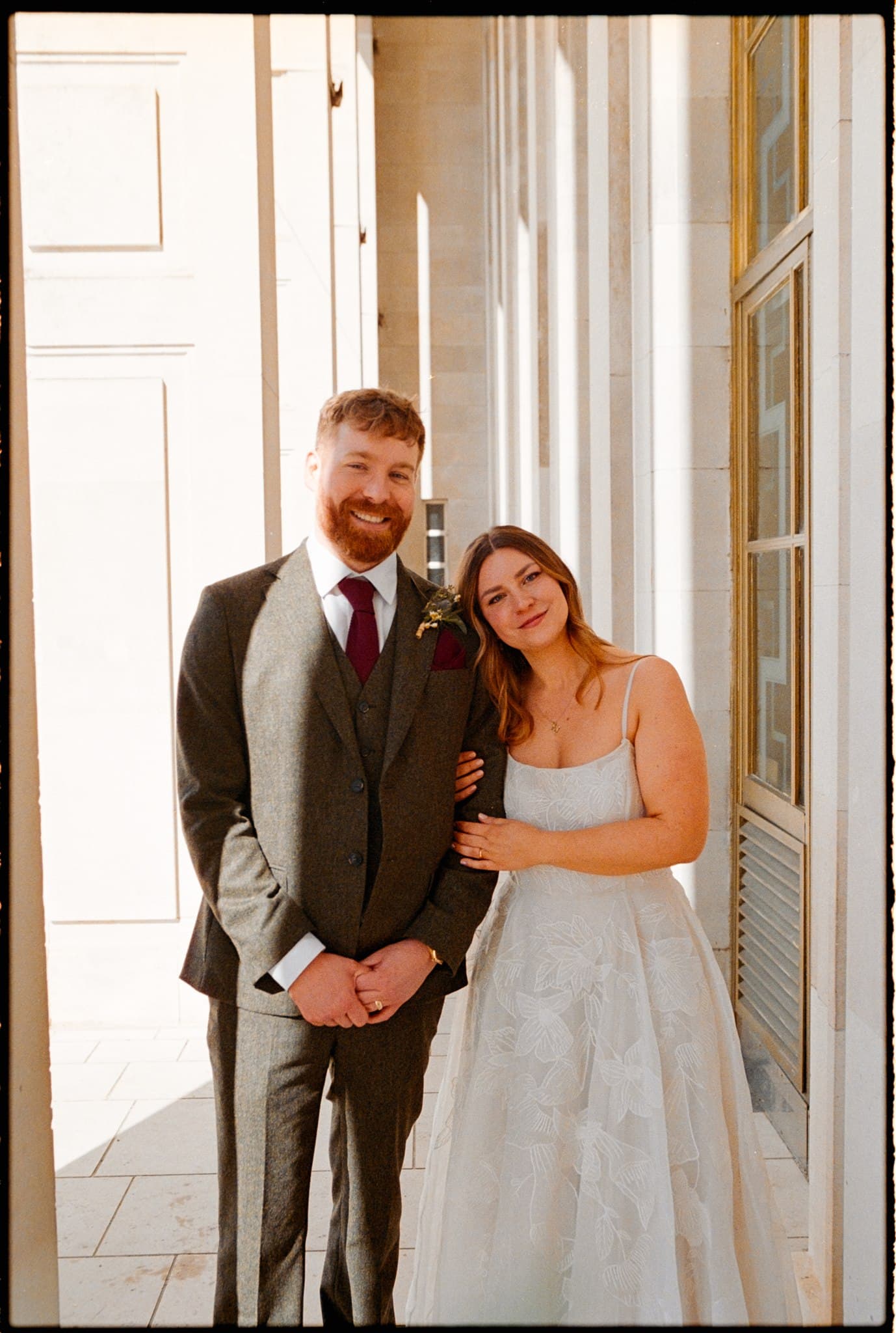 A couple in wedding attire standing close together in a sunlit doorway. film photo