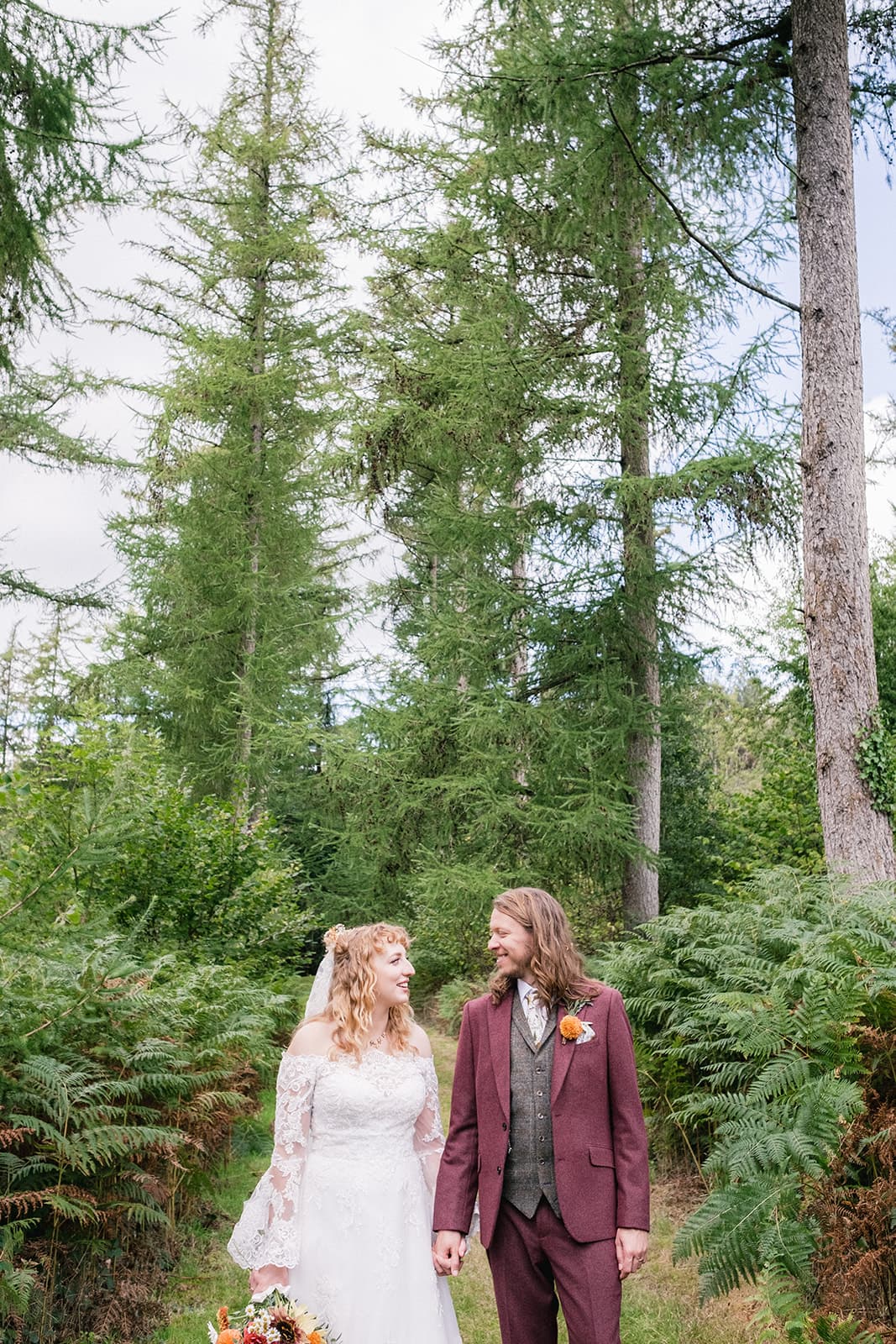 A bride in a white dress and a groom in a maroon suit standing in a forest.