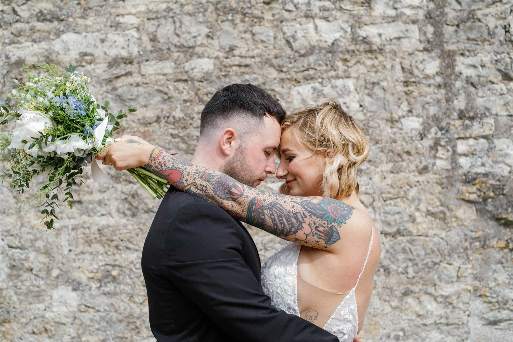 A bride and groom share a loving embrace in front of a rustic stone wall, captured beautifully by Ivy and Pine Photography.