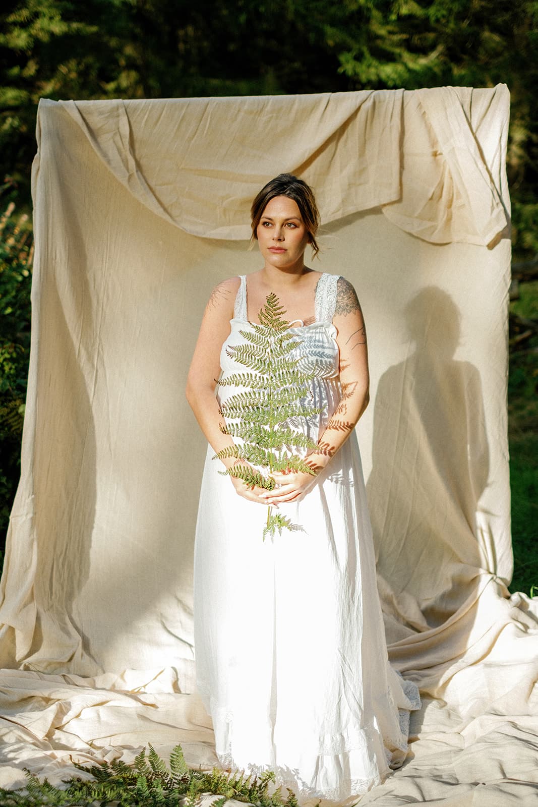 A woman in a white dress stands in front of a cream backdrop outdoors, holding a fern. Sunlight filters through trees, casting soft shadows. Serene atmosphere.