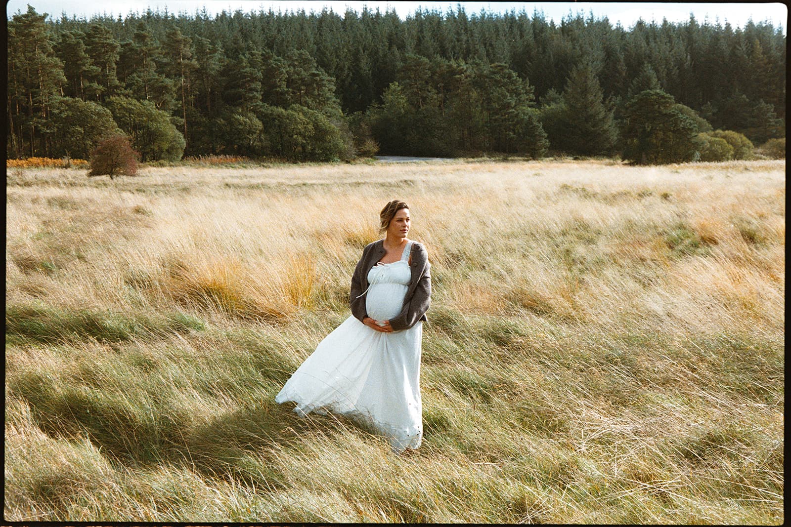 Pregnant woman in a white dress and gray cardigan stands in a sunlit grassy meadow. Forest in the background, atmosphere is serene and reflective.