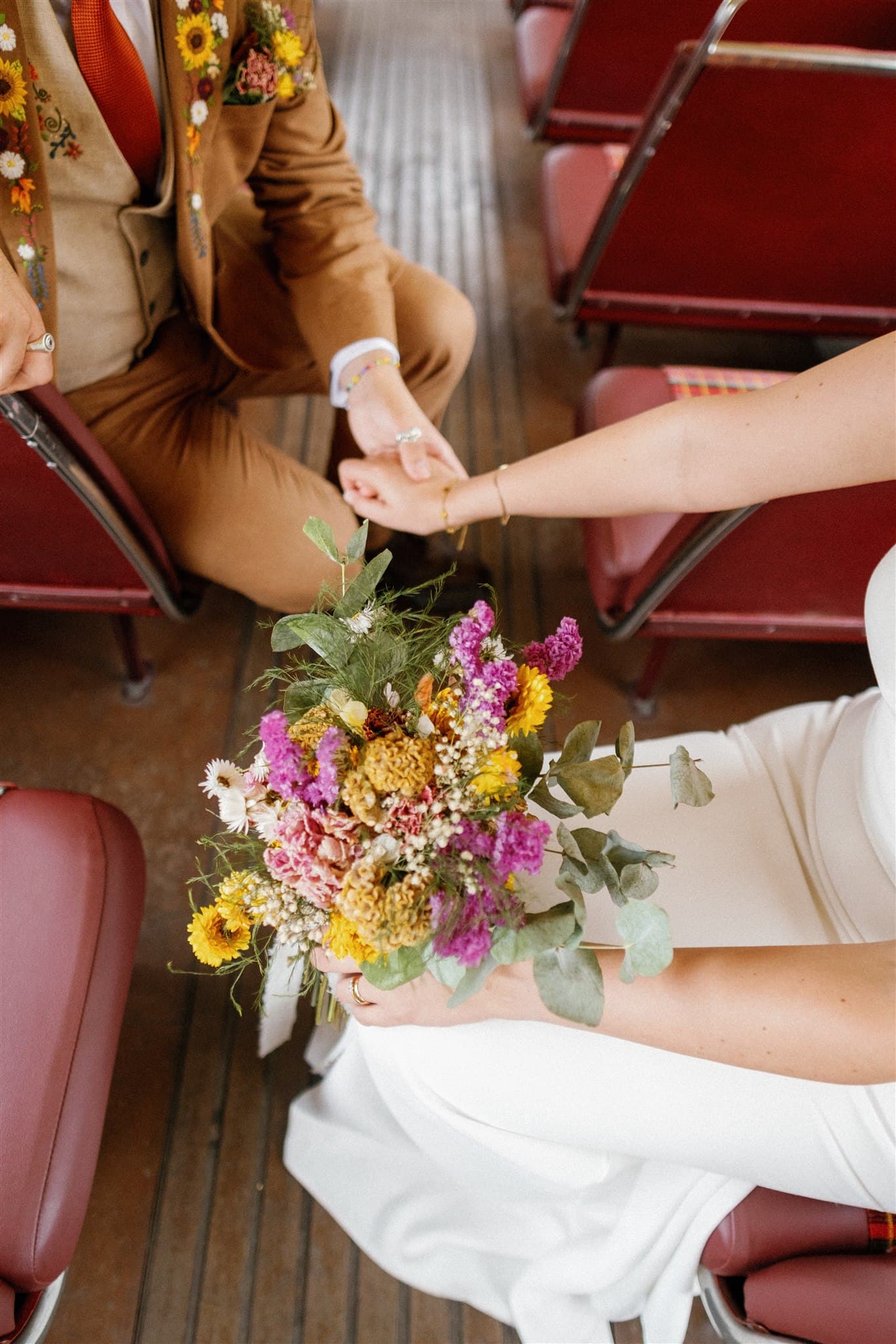 A couple in wedding attire holds hands on a bus; the bride's bouquet features colorful wildflowers. The setting conveys a vintage, joyful ambiance.