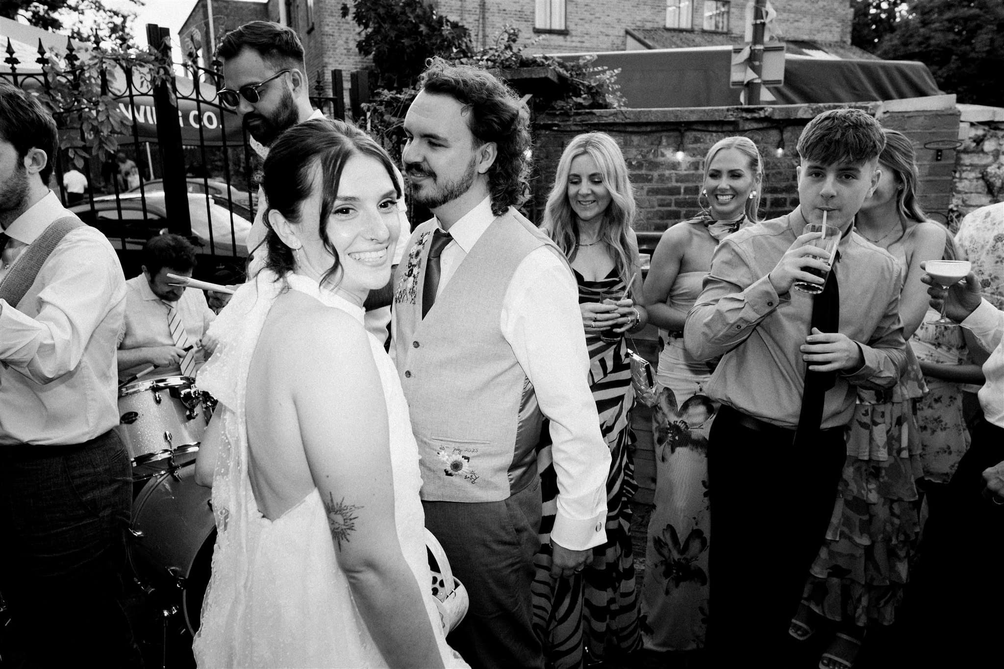 A joyful bride smiling at the camera, surrounded by guests in formal attire at an outdoor celebration. The atmosphere is lively and festive. Black and white.