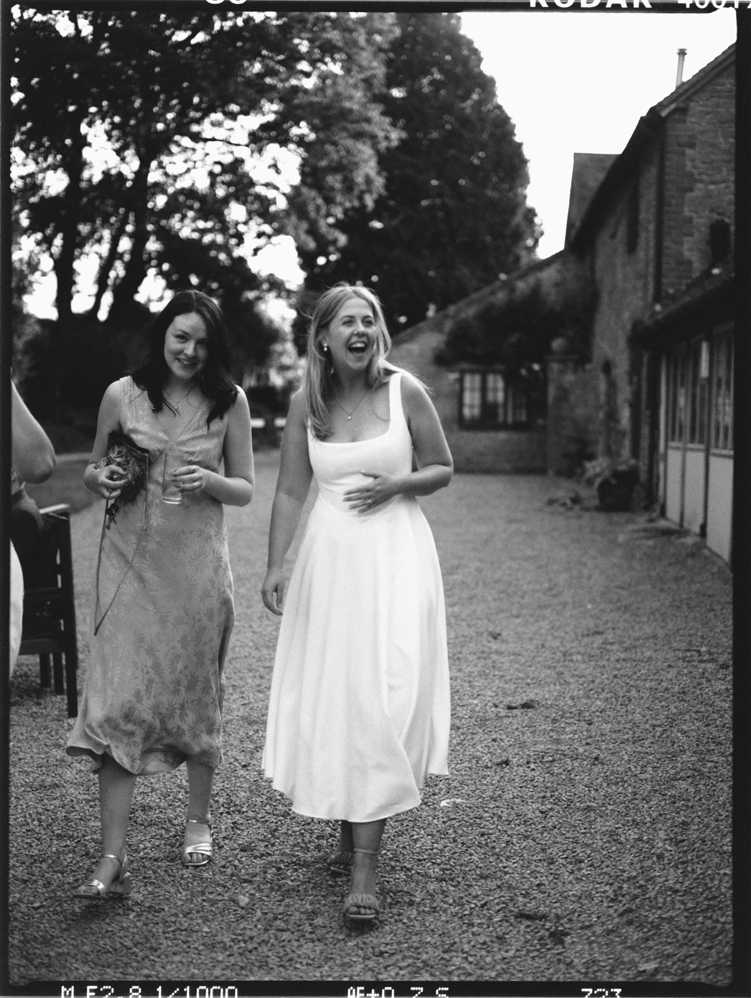 Two women in dresses, one in light and the other in dark, walk on a gravel path between trees and rustic buildings, sharing a joyful laugh.